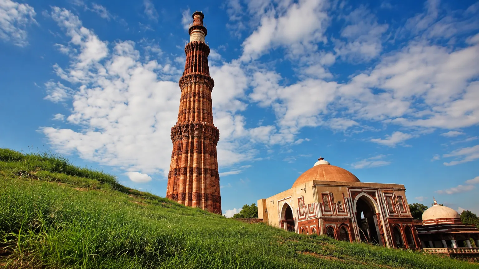 Qutub Minar rising above the historic complex in Delhi under a bright blue sky with surrounding Mughal architecture.