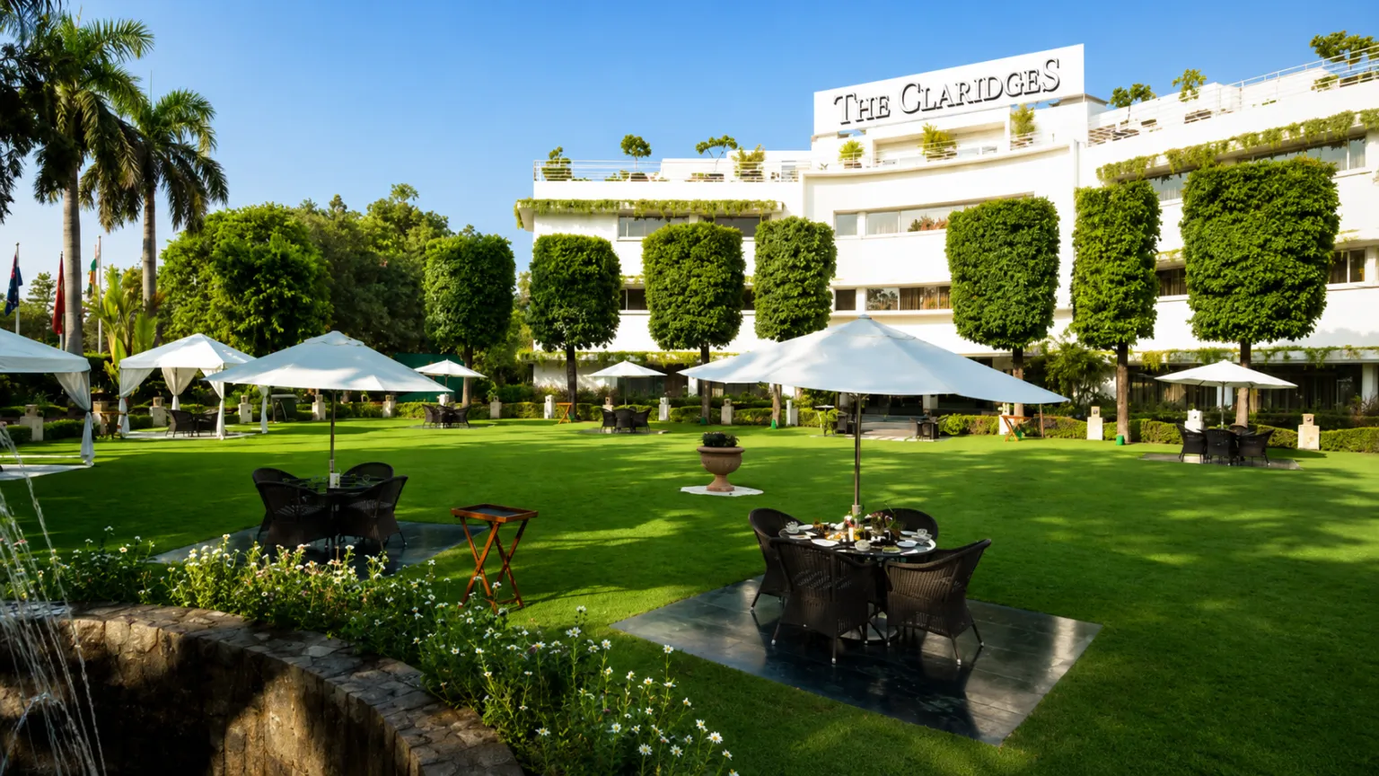 Beautiful gardens at The Claridges New Delhi featuring manicured lawns, outdoor seating, white umbrellas, and luxury hotel surroundings.