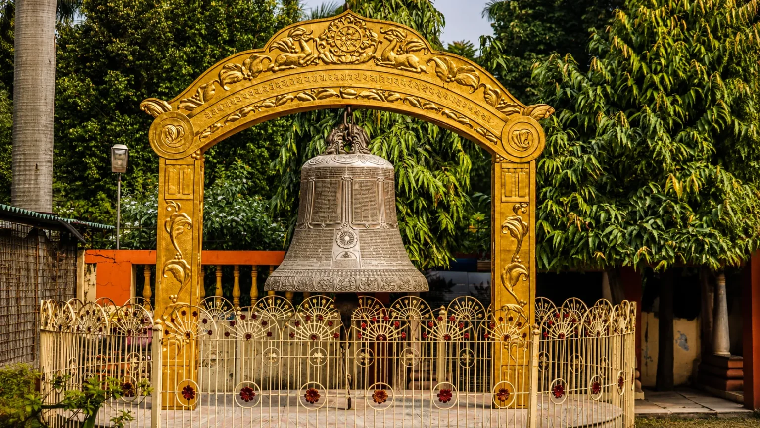 Historic Ashoka Bell displayed beneath an ornate golden arch inside the Sarnath archaeological complex surrounded by lush greenery.