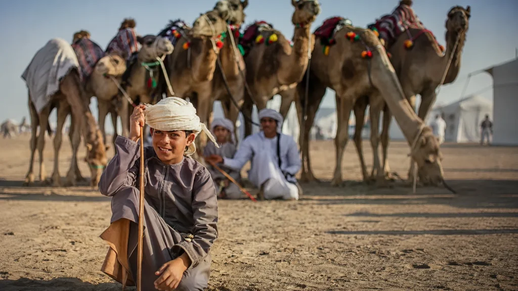 Young boy in traditional attire sitting on desert sand with decorated camels and local camel handlers in the background during a camel safari experience in Rajasthan, India.