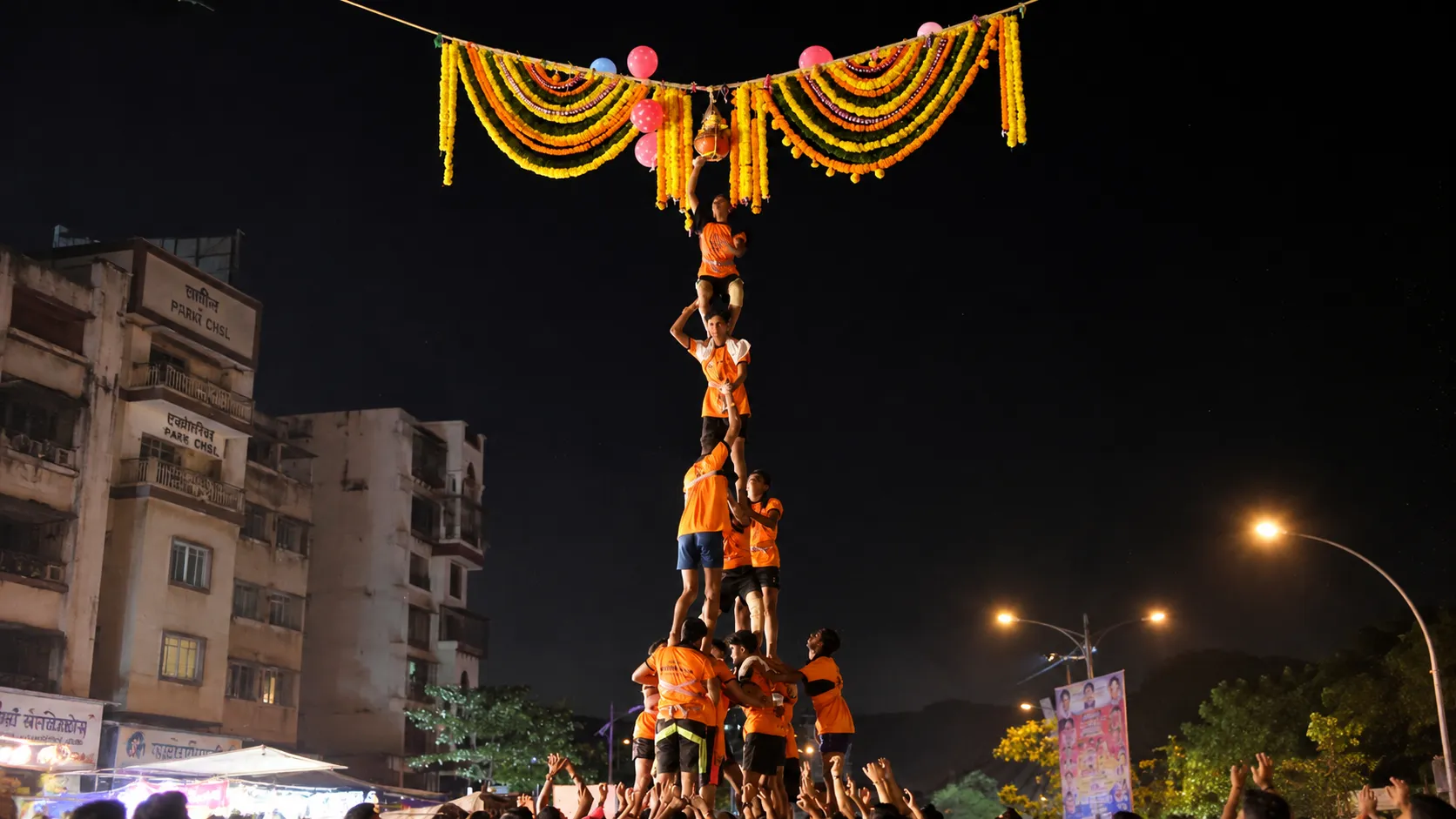 A human pyramid formed by devotees during the Dahi Handi festival on Janmashtami, reaching for the decorated handi suspended above amid colorful floral decorations and cheering crowds at night.