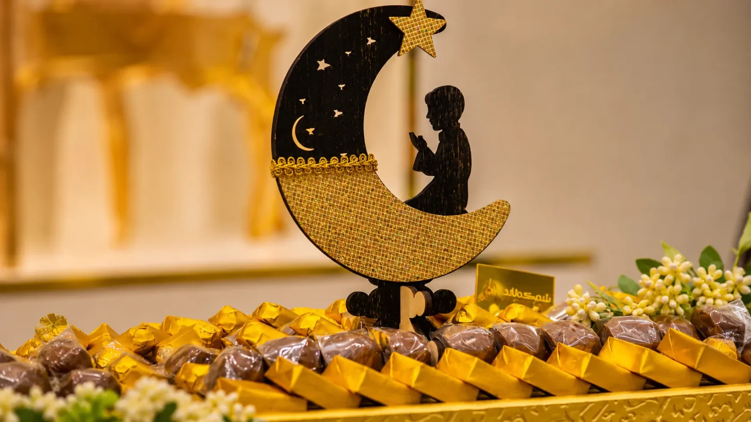 Decorative crescent moon with a praying child silhouette displayed among assorted chocolates and sweets for an Eid celebration.