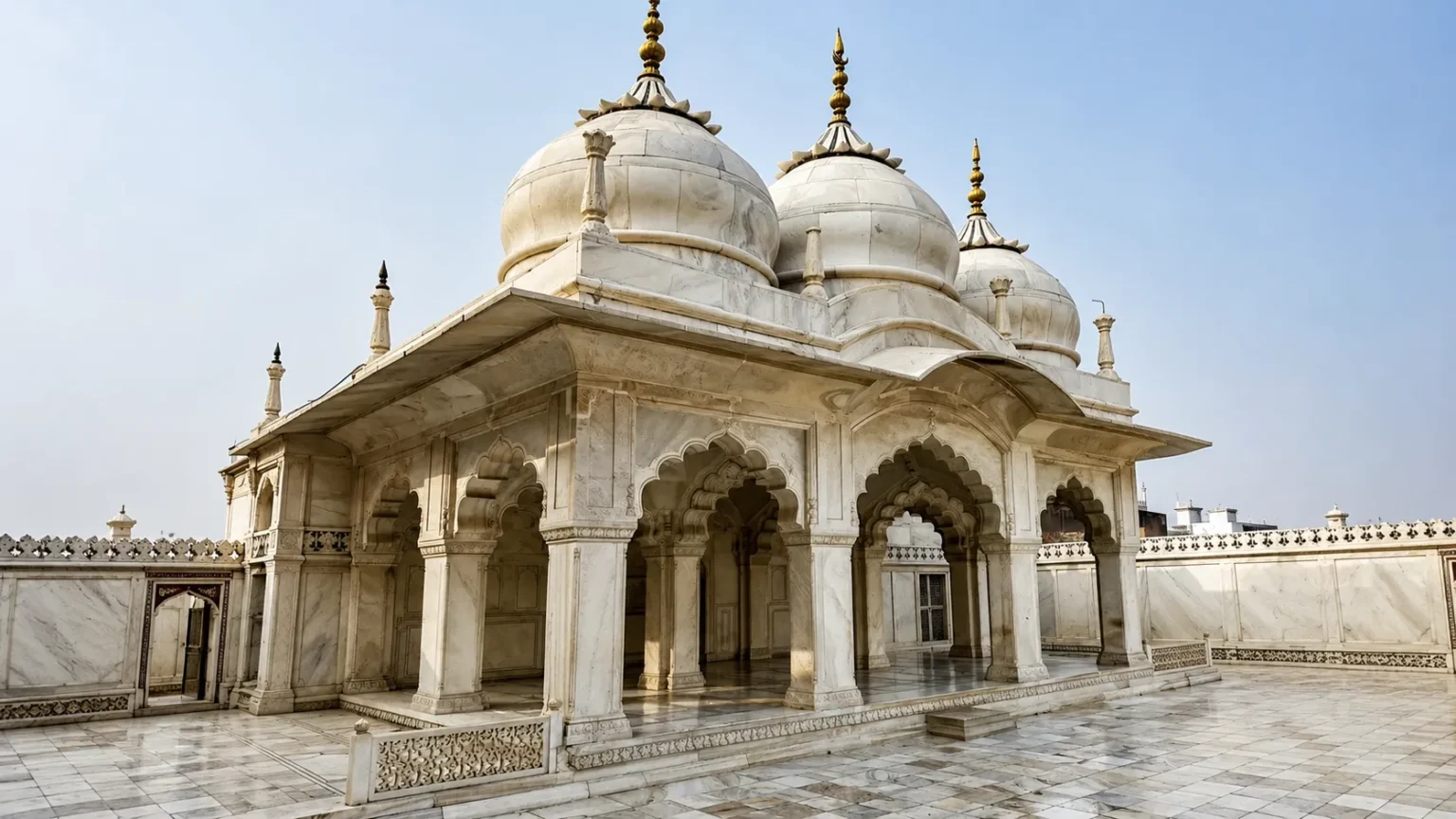 White marble pavilion featuring domes, ornate arches, carved pillars, and polished marble flooring under a clear blue sky.