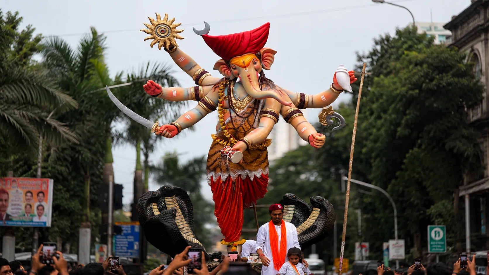 A magnificent Lord Ganesha idol carried during a Ganesh Chaturthi procession, surrounded by devotees on a city street with vibrant decorations and traditional celebrations.
