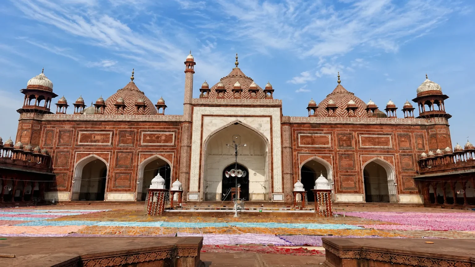 Red sandstone façade of Jama Masjid in Agra with elegant domes, arched entrances, and intricate Mughal architecture.