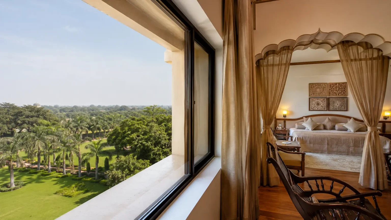 A luxurious hotel suite with a traditional Mughal arched canopy over the bed looking out onto a sprawling green lawn view in Agra.