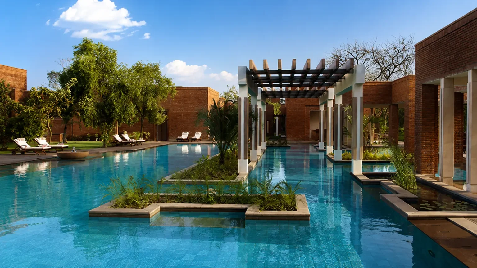 A sprawling luxury outdoor swimming pool with red brick pavilions and lush trees at ITC Mughal resort in Agra.