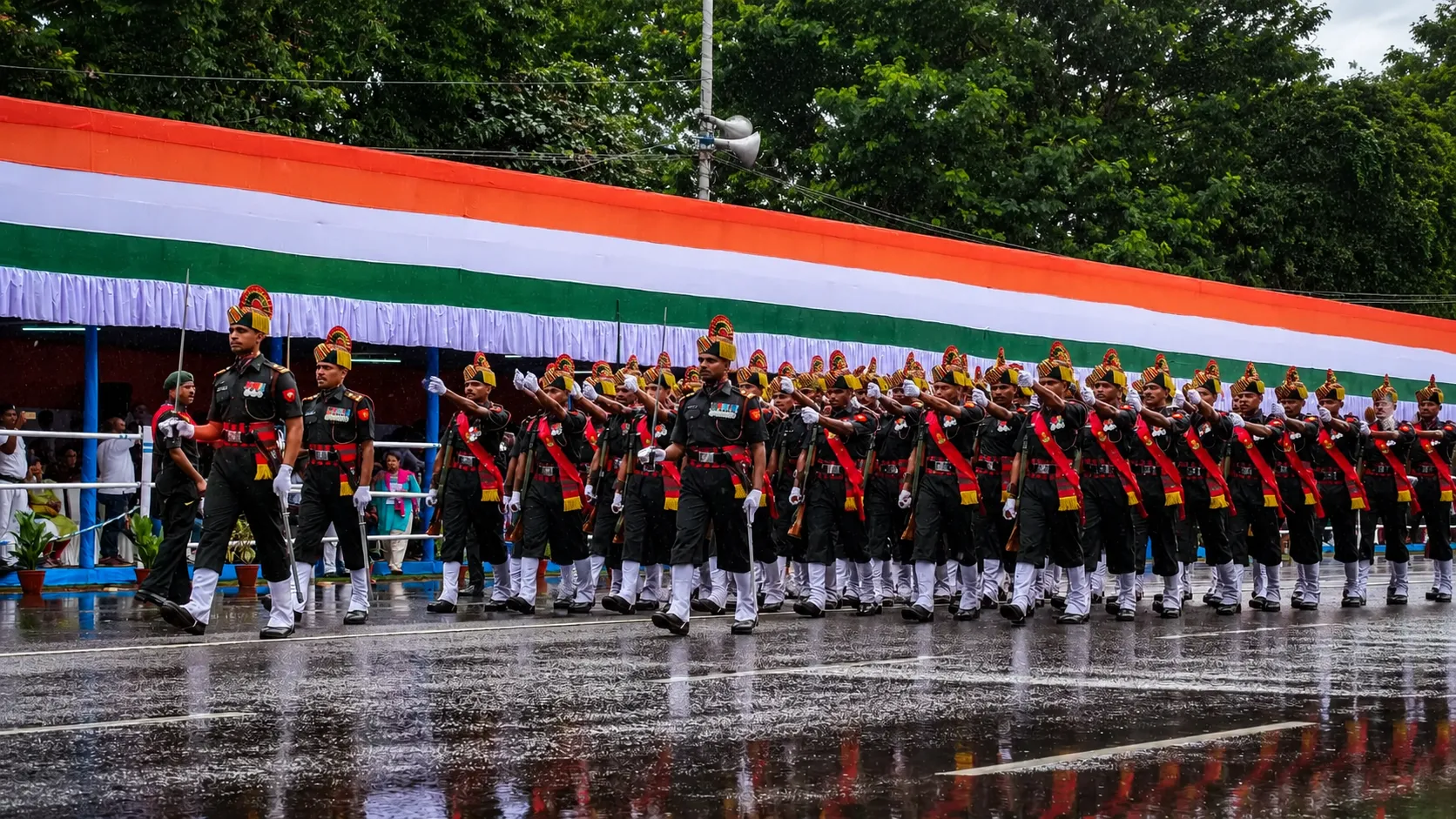 Indian Army soldiers marching in formation during a rainy ceremonial parade with the Indian tricolour backdrop and reflections on the wet road.