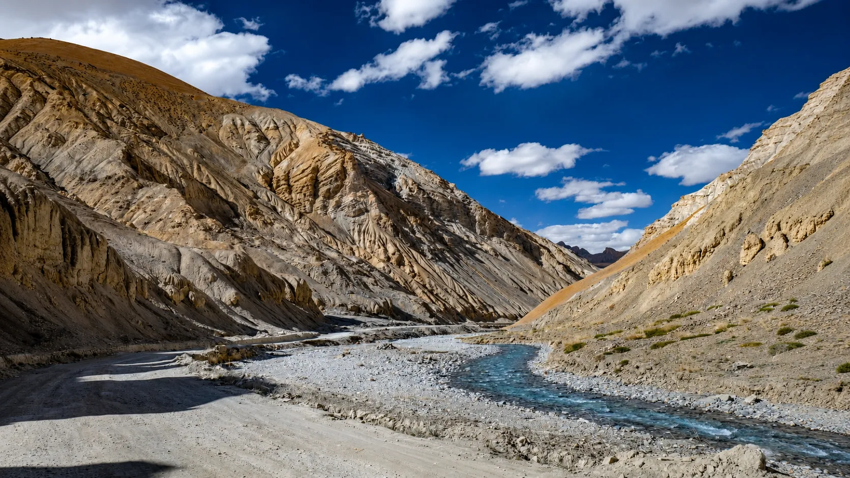 A scenic mountain valley in Ladakh featuring rugged rocky cliffs, a winding crystal-clear stream, and a dramatic blue sky with white clouds in the Indian Himalayas.