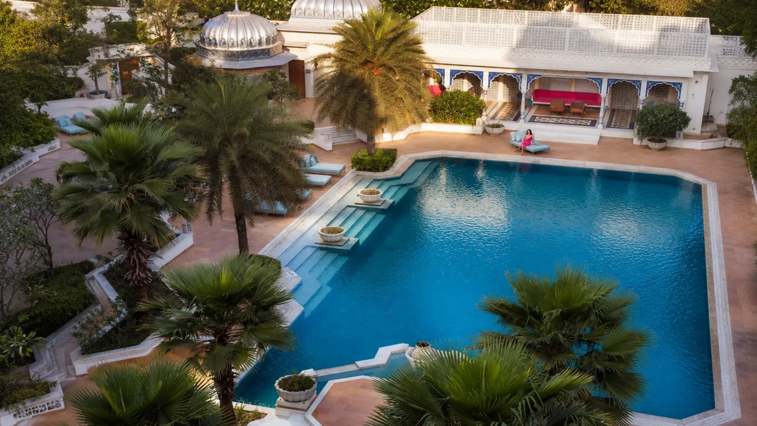 Serene swimming pool at The Leela Palace Udaipur surrounded by lush palm trees, elegant architecture, and tranquil luxury overlooking Lake Pichola.