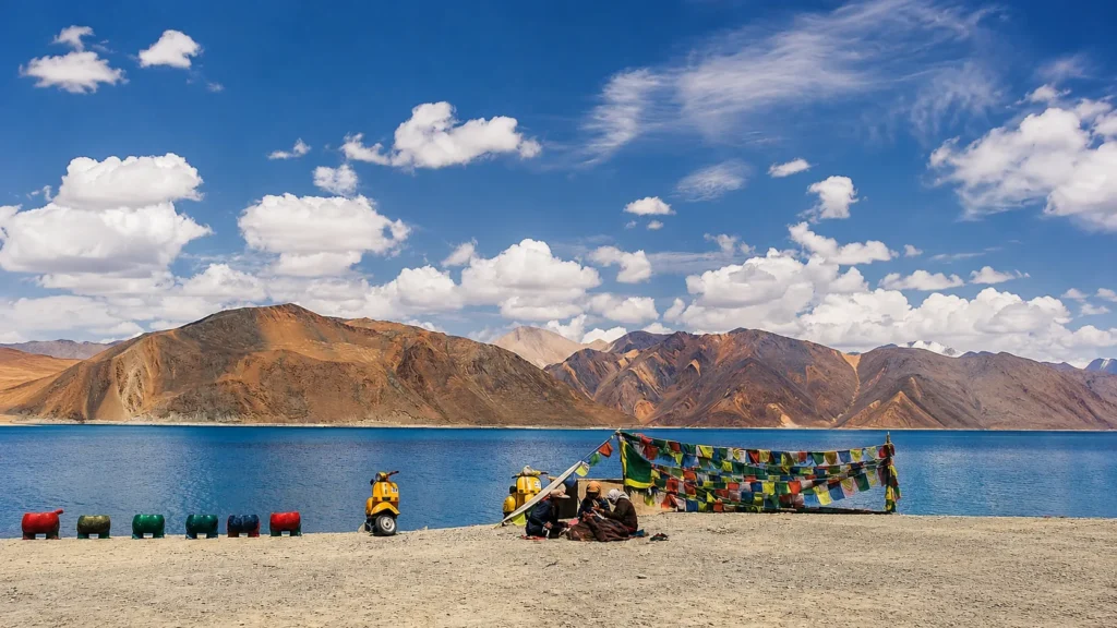 Scenic view of Pangong Lake with colorful prayer flags, rugged Himalayan mountains, and a bright blue sky in Ladakh, India.