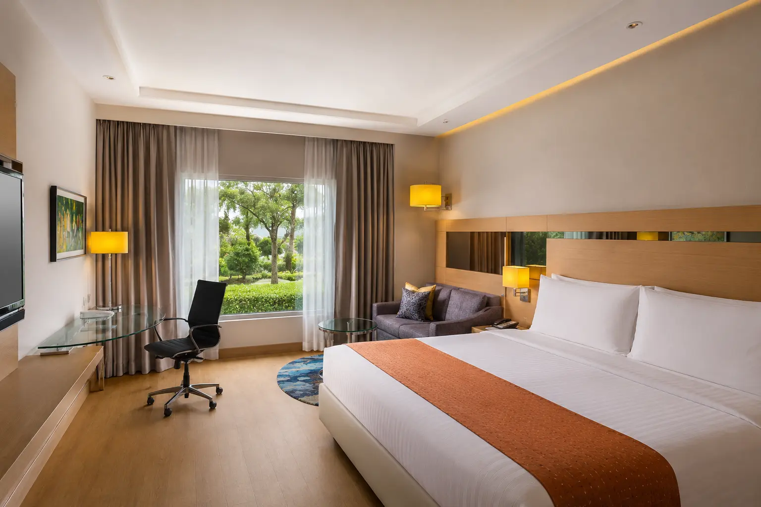 A modern hotel bedroom with a large window overlooking green gardens, a glass work desk, and a king bed at Radisson Hotel Agra.