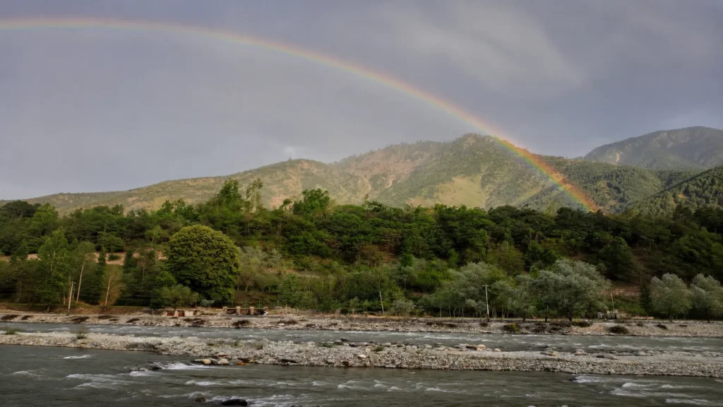 A vibrant rainbow stretching across forested Himalayan mountains above a flowing river under dramatic post-rain clouds.