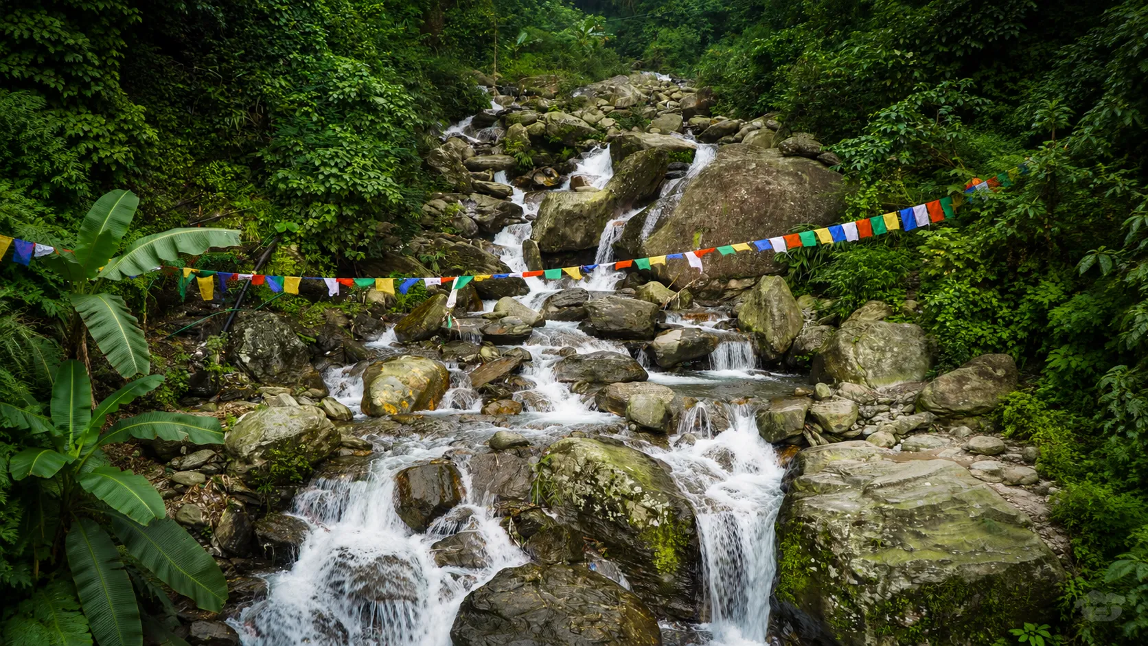 A cascading mountain waterfall flowing over moss-covered rocks through a dense green forest with colorful prayer flags creating a peaceful Himalayan landscape.