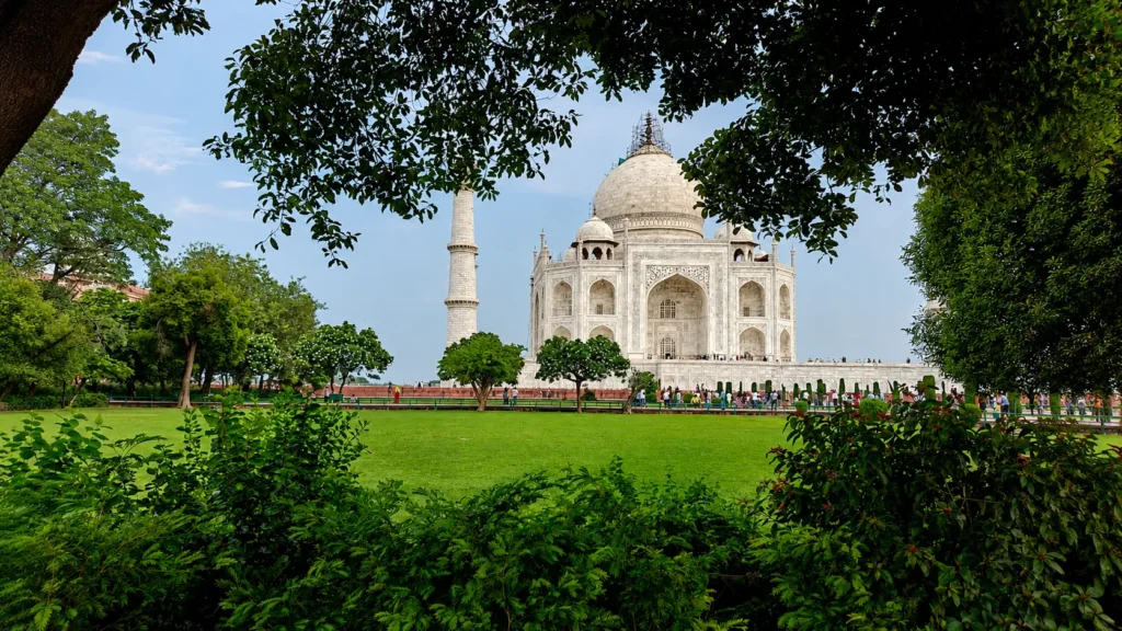 The Taj Mahal viewed through lush green trees and gardens on a clear day, showcasing its iconic white marble architecture in Agra, India.
