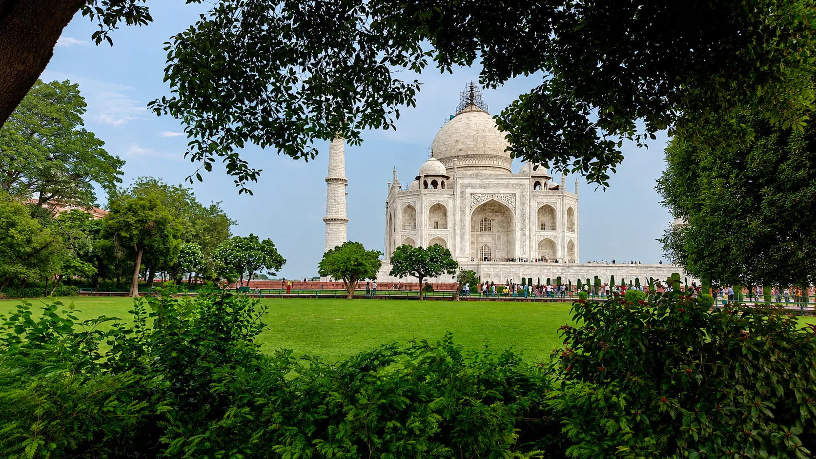 The Taj Mahal viewed through lush green trees and gardens on a clear day, showcasing its iconic white marble architecture in Agra, India.