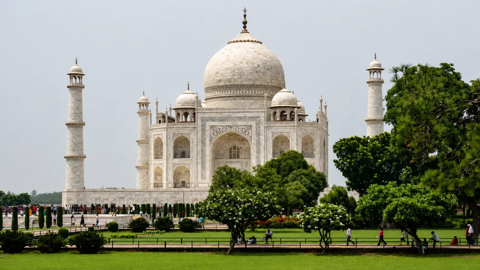 Panoramic view of the Taj Mahal with lush green gardens, flowering trees, and visitors exploring the UNESCO World Heritage Site in Agra.