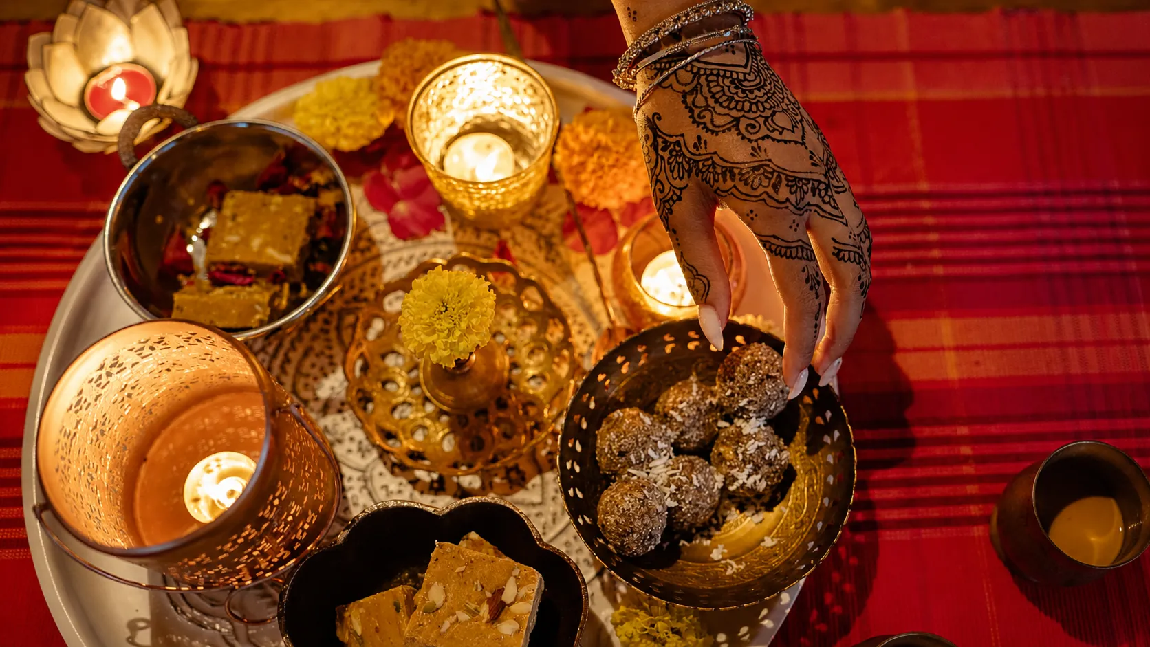 A beautifully arranged Diwali festive platter featuring traditional Indian sweets, glowing diyas, marigold flowers, and a hand adorned with intricate mehndi reaching for a sweet during the celebration.