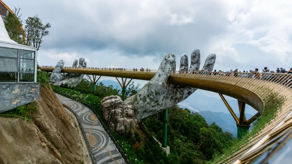 Wide-angle view of the Golden Bridge at Ba Na Hills in Da Nang, Vietnam, supported by giant stone hands under a dramatic evening sky with warm golden light and lush mountain scenery.