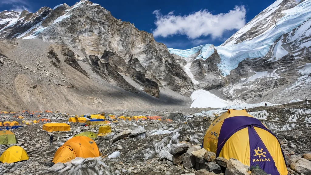 High-altitude mountain base camp with colorful tents scattered across rocky terrain beneath a massive snow-covered Himalayan peak and glacier.