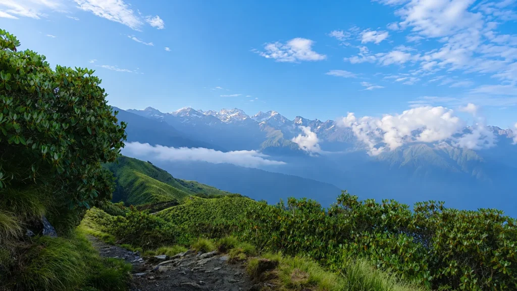 Scenic Himalayan mountain landscape with a rocky hiking trail, lush green hills, mist, and snow-capped peaks under a blue sky.