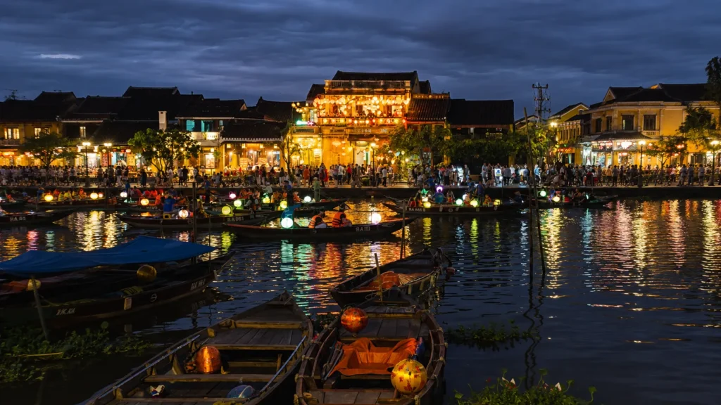 Traditional lantern boats floating on the Thu Bon River with illuminated heritage buildings and vibrant evening lights in Hoi An Ancient Town, Vietnam.