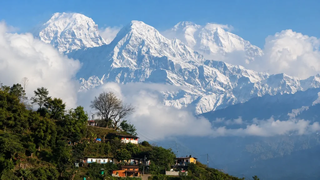 Snow-covered Himalayan mountains rising above a green hillside village beneath a clear blue sky
