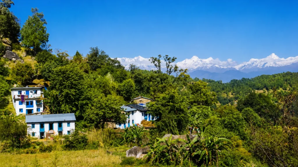 Picturesque Himalayan village with white and blue houses surrounded by lush greenery and snow-capped mountains in the background.