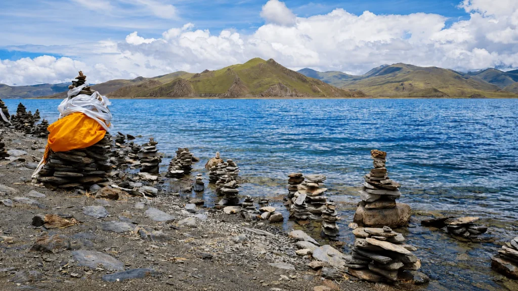 Serene high-altitude lake with stacked stone cairns along the rocky shoreline, colorful prayer cloth, and green mountain ranges beneath a cloudy blue sky.