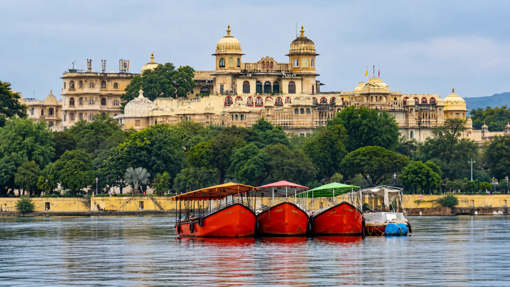 Colorful boats floating on Lake Pichola with the ornate City Palace of Udaipur and lush green trees in the background under a soft cloudy sky.