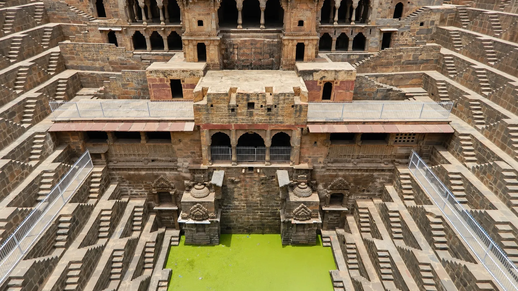 Ancient Chand Baori stepwell in Rajasthan featuring symmetrical stone steps, ornate architecture, and a green water pool.