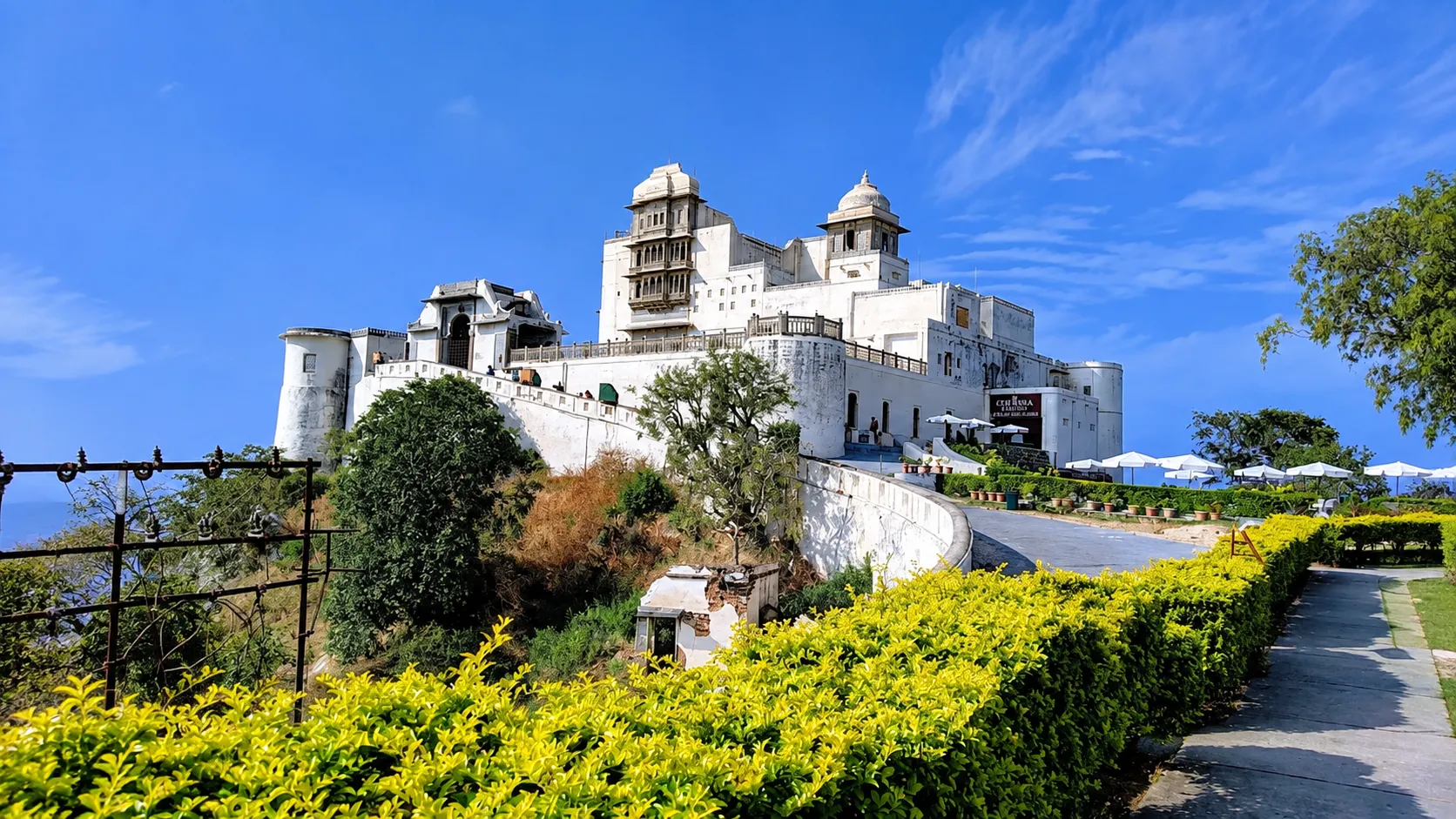 White hilltop palace with ornate domes and traditional Rajasthani architecture, surrounded by landscaped gardens and bright green hedges under a clear blue sky.