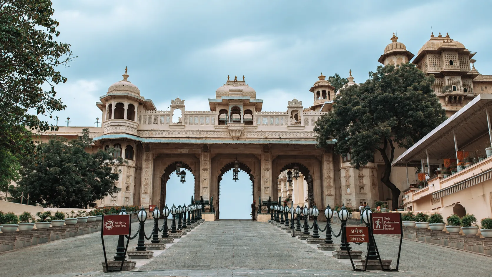 Ornate historic palace entrance in Jaipur with three carved arches, traditional domes, decorative architecture, trees, and a paved walkway lined with black lantern posts.