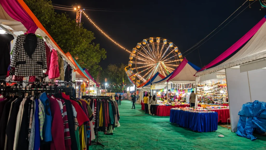 Colorful Dussehra fair at night with decorated market stalls, clothing vendors, a brightly lit Ferris wheel, and visitors