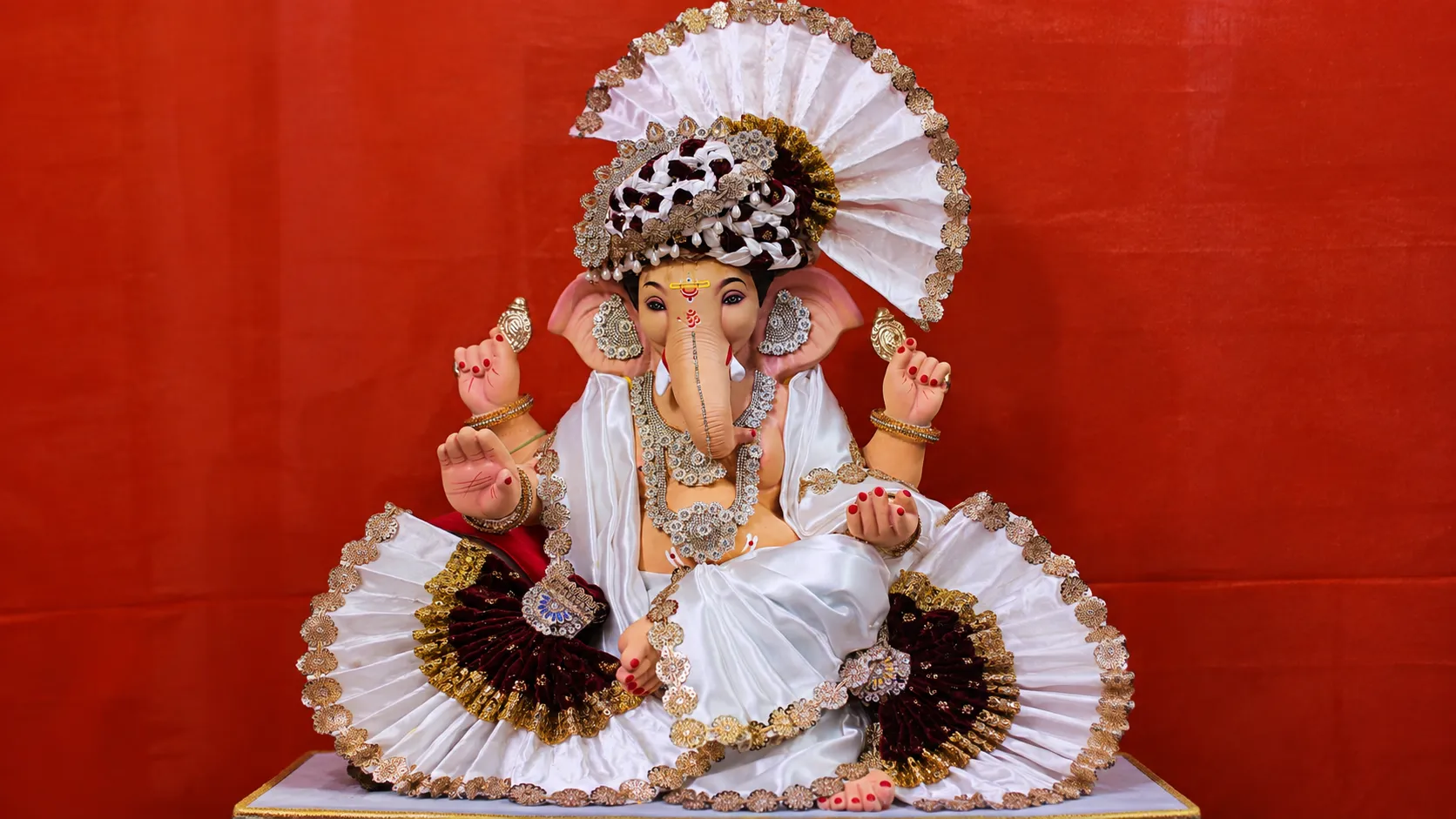 Lord Ganesha idol dressed in white silk and gold-trimmed decorations against a red backdrop