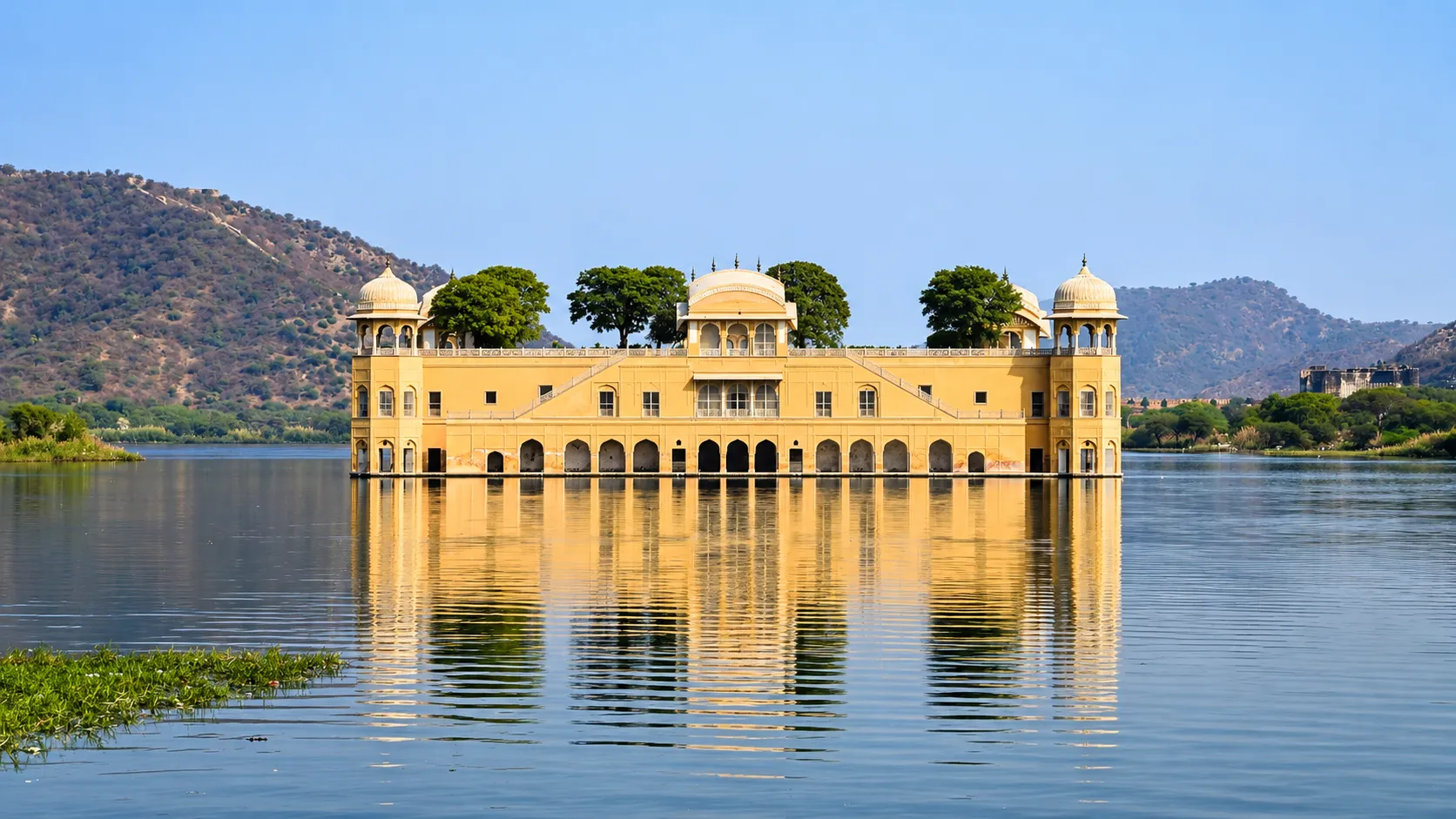 Jal Mahal palace standing in the middle of Man Sagar Lake in Jaipur, surrounded by hills, trees, and calm reflective water under a clear blue sky.