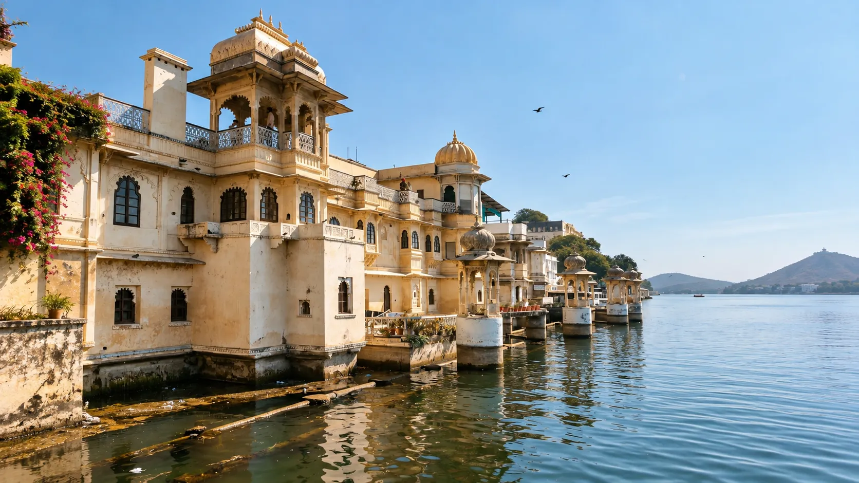 Historic Rajasthani palace architecture beside a calm lake in Udaipur, with ornate domed pavilions, carved balconies, water reflections, distant hills, and a clear blue sky.
