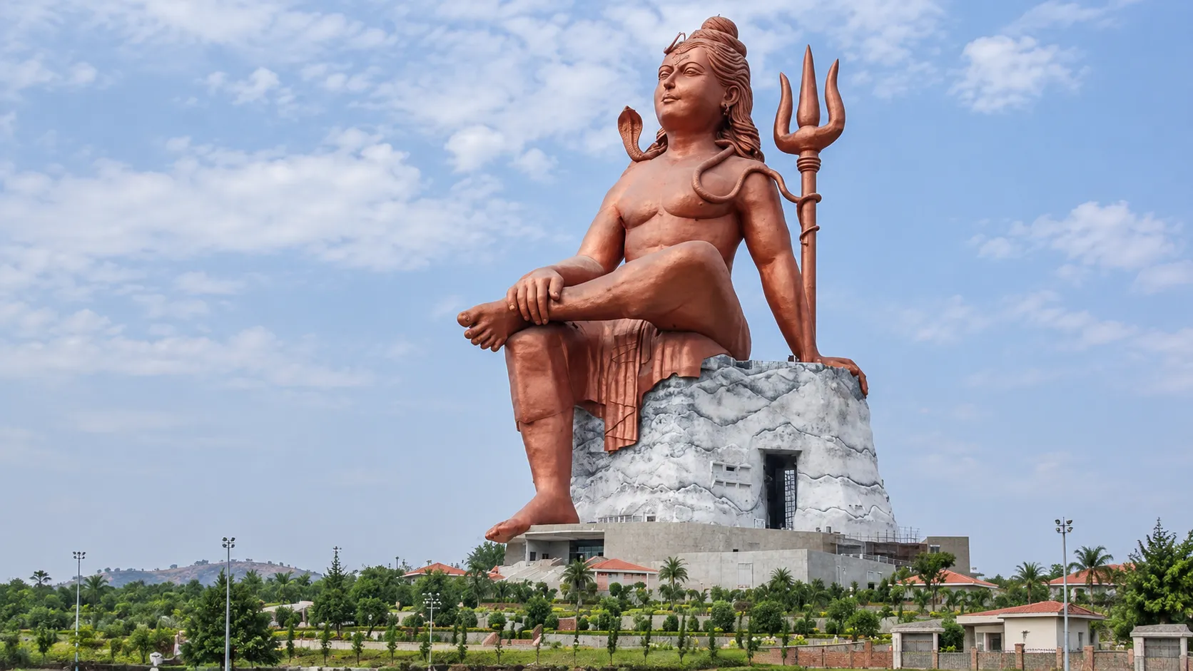 Giant seated Lord Shiva statue with a trident overlooking the landscaped temple complex under a blue sky