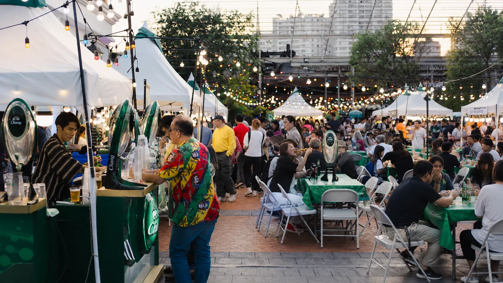 Crowded outdoor beer festival with food stalls, string lights, dining tables, and festive evening atmosphere