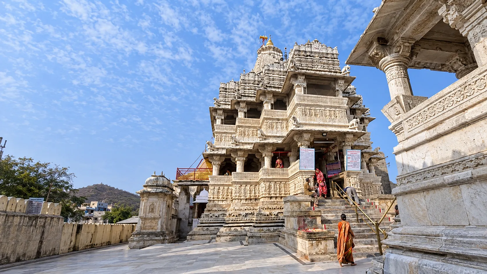 Intricately carved white marble Jain temple with ornate pillars, domes, and detailed stonework beneath a bright blue sky, with visitors approaching the entrance.