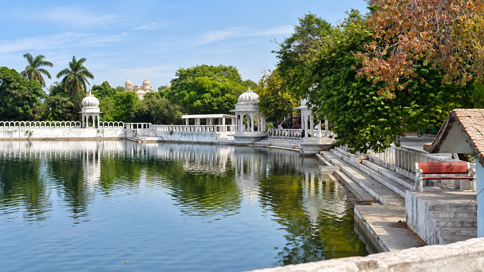 A serene heritage pond surrounded by lush green trees, white domed pavilions, ornate railings, stone steps, and a red bench under a clear blue sky.
