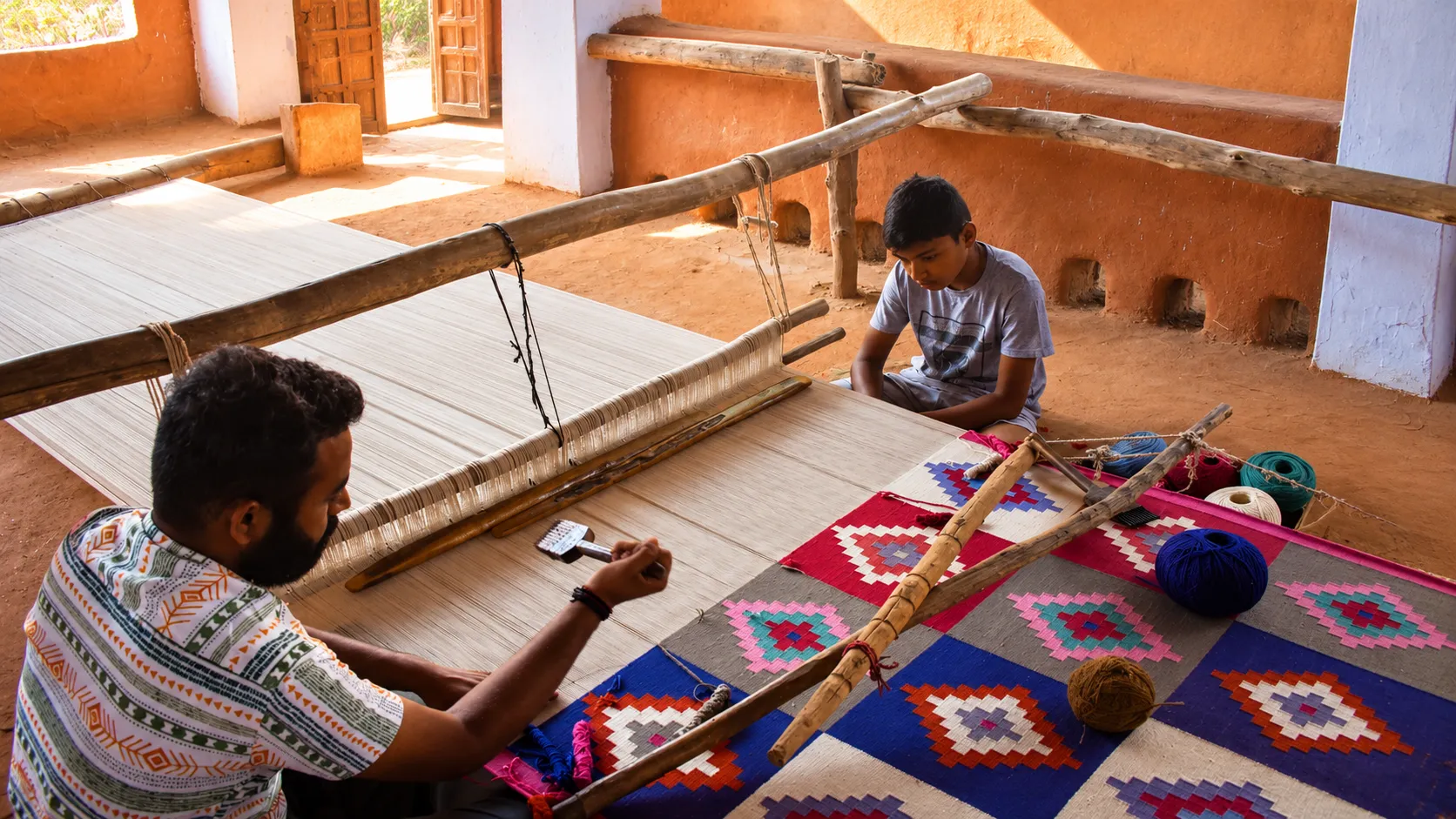 Carpet artisan carefully repairing or weaving a patterned rug inside a traditional shop filled with stacked carpets