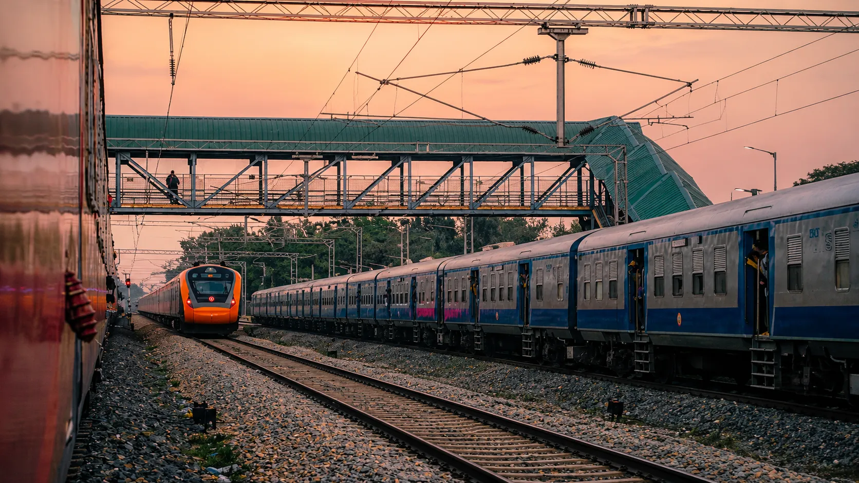 Blue passenger train and orange train on parallel railway tracks beneath a station footbridge at sunset.