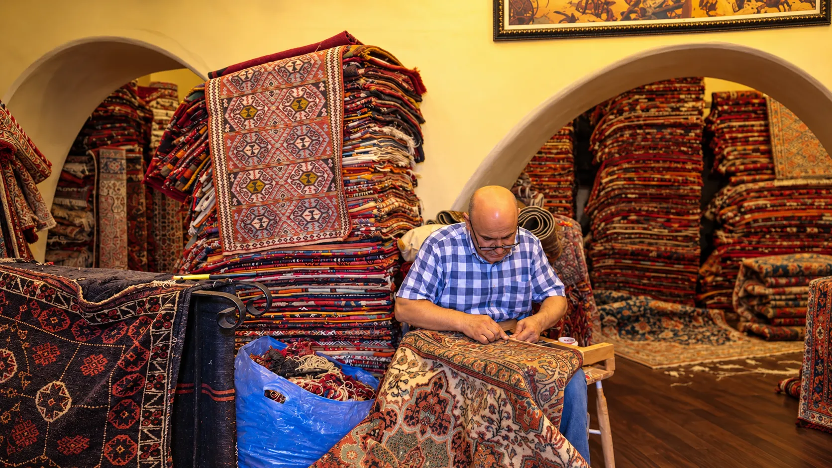 Artisan working on a patterned carpet inside a traditional rug shop filled with colourful rugs stacked beneath arched alcoves
