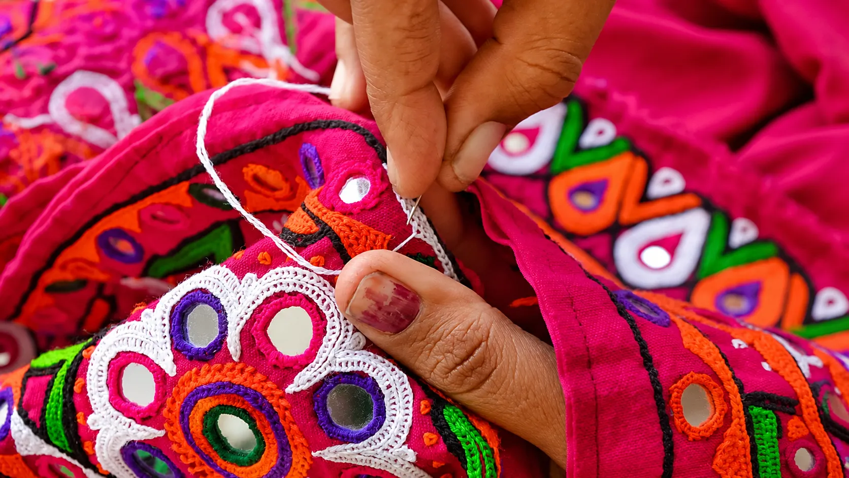 Close-up of hands stitching colourful Gujarati embroidery with mirror work and vibrant pink, orange, green, and purple threads