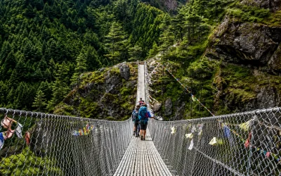Hikers crossing a suspension bridge through a lush Himalayan mountain forest with rocky cliffs and colorful prayer flags